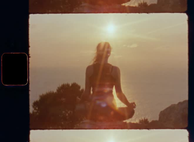 The backside of a woman sitting on a rock ledge overlooking the sun over the ocean while she meditates and her long hair sways in the wind as she connects with herself and nature in an epic and quiet setting without distractions