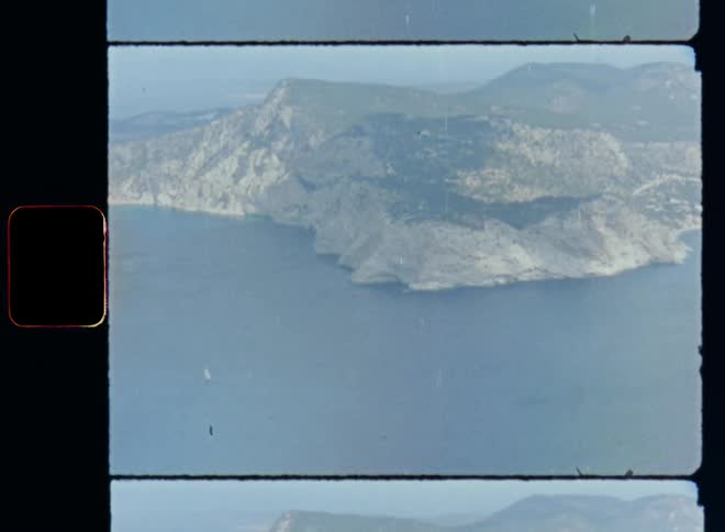 Aerial drone footage of an island in the ocean or sea with high mountains and steep cliffs covered in green forests and grassland surrounded by shore and coastline and a white sailing boat is floating in the water on Super 8mm film footage