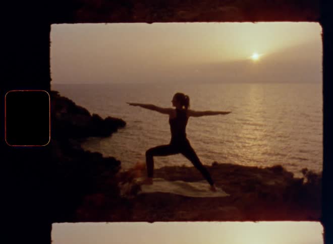 Handheld dynamic 8mm film footage of the silhouette of a female sportive woman doing yoga next to the ocean at dusk embodying strength and power with a healthy and trained body as the sun sets behind a cloud wall at the horizon over the ocean
