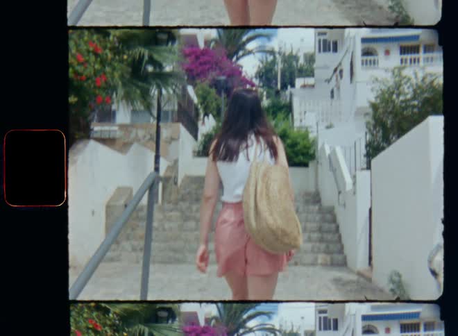 Lightly dressed woman in summer clothes carrying a handbag walks up a set of stairs outdoors between some residential buildings or apartments with a palm tree and colourful flowering bushes and plants in private gardens behind walls and fences