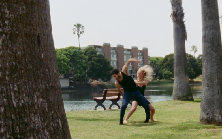 Two dancers dance together in park across water from apartment building