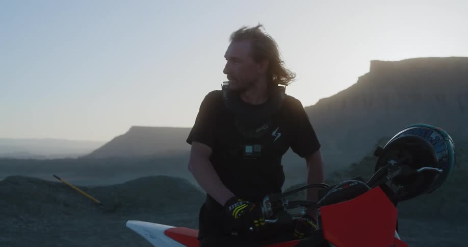A man sits on a motorcycle and looks around in a desert at dawn