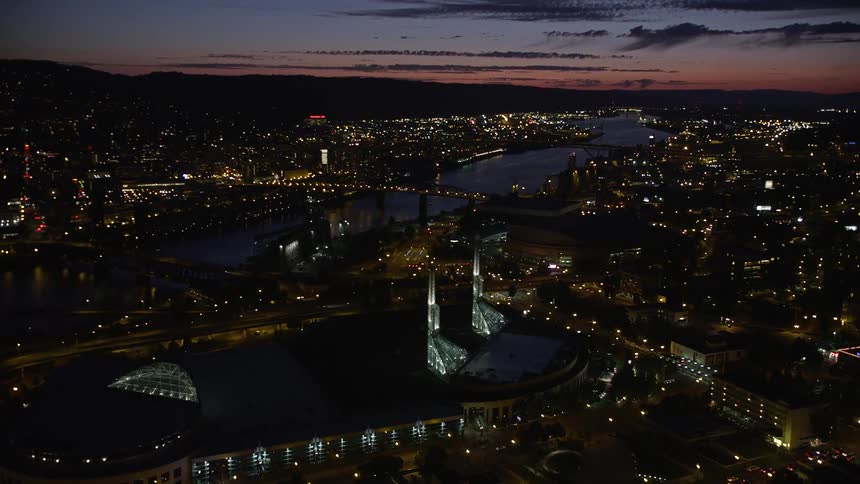 Aerial footage at night of Portland's cityscape as seen from above the Willamette River approaching the Oregon Convention Center and Broadway Bridge in the Lloyd District