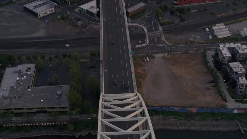 Aerial footage of vehicles driving over the arched Fremont Bridge in downtown Portland Oregon during a summer sunset with partly cloudy skies and the American flag waving in the wind