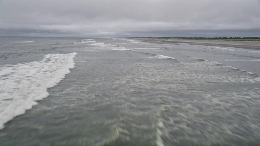 Low flying aerial footage showcases the beautiful coastline of Ilwaco Washington during the summer with crashing waves and an empty beach below