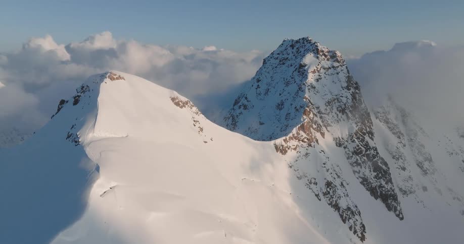 A rocky mountain range is covered in snow and the peak is at the height of the nearby clouds