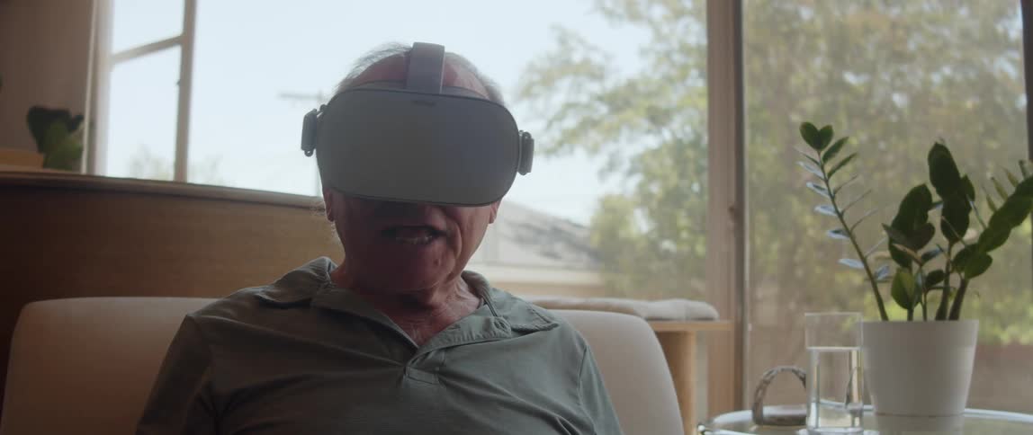 An elderly man sits indoors while enjoying wearing a virtual reality headset