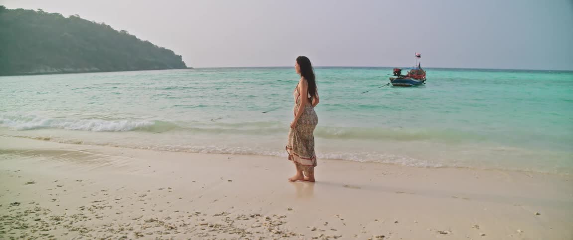 Woman walking on the beach sand next to a sea with crystalclear water wearing a stylish long dress