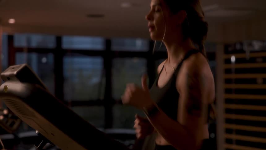 Close up of a woman running on a treadmill indoors during the day in a dim lit room