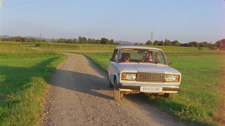 Man drives car onto dirt road with farm equipment operating in crop field in the background