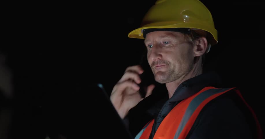 Close up of a man wearing a hardhat and caution vest while talking on his phone looking at a tablet at night