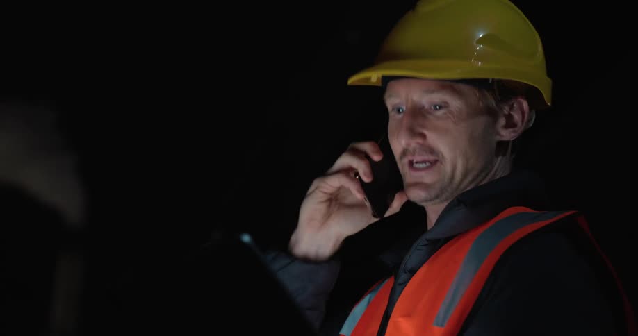 A man in a construction hat is talking with a cell phone to his ear while looking at a laptop