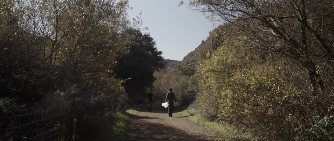 Two men walk along a dirt path surrounded by trees