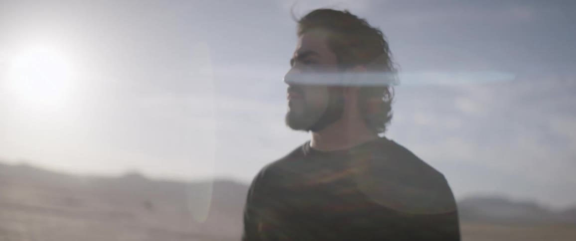 Dark haired man with facial hair stands on desert plain
