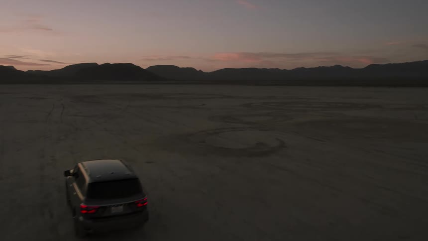 Drone view of an suv driving over the desert sand at dusk with mountains on the horizon