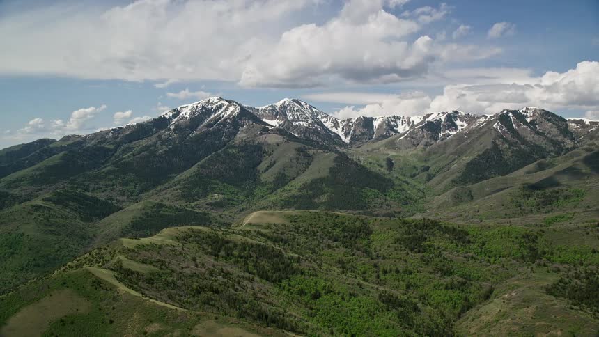 Aerial footage flying by snowy peaks and green ridges of Oquirrh Mountains in Utah during summer