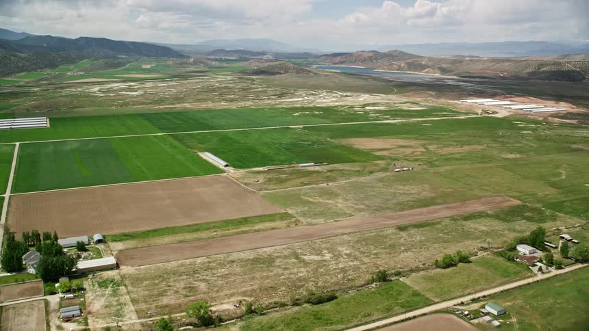 Aerial footage of a vast agricultural area with a lake and mountain range on the horizon captured during a cloudy day in Utah's countryside near Gunnison Reservoir