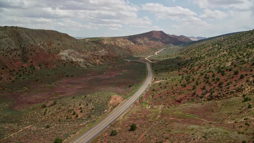Aerial footage tracking a white car on Highway 89 in Glen Canyon National Recreation Area Utah Arizona as it passes through large hills in the desert with lush vegetation on a partly cloudy day during summer