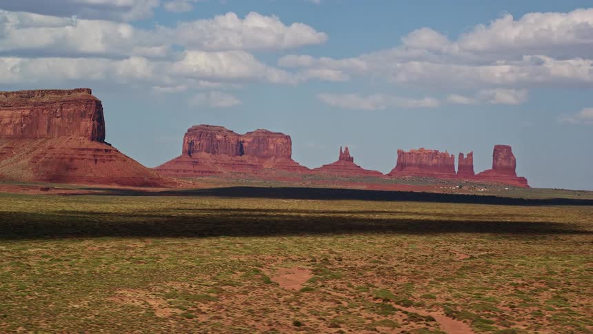  aerial footage showing the passing of four desert buttes in Monument Valley Arizona and Utah with partly cloudy skies and a view of the surrounding landscape during daytime