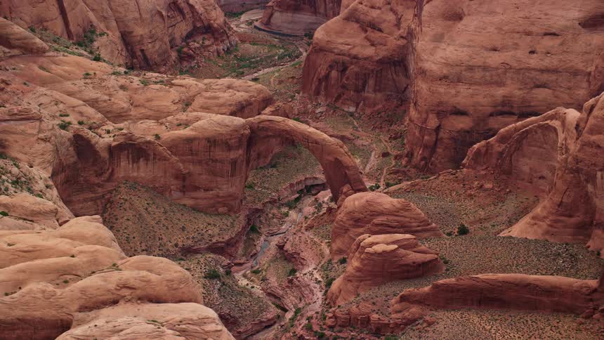 aerial footage capturing the stunning natural stone arch of Rainbow Bridge National Monument situated in an arid desert landscape with partly cloudy skies showcasing the unique rock formations and the beauty of the Southwest United States during the summ