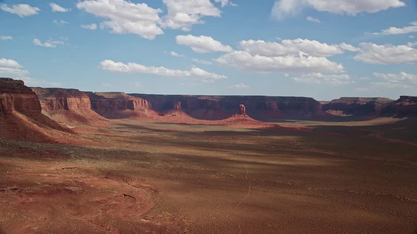 Approaching a flat-topped butte in a wide valley surrounded by mesas on the Navajo Nation Reservation in Arizona and Utah captured in aerial footage during a partly cloudy day in the Southwest United States