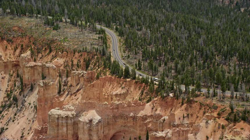 Aerial footage of Highway 63 surrounded by forest on the edge of a canyon cliff in Bryce Canyon National Park Utah showcasing the famous Pink Cliffs and natural rock formations with light traffic and partly cloudy skies