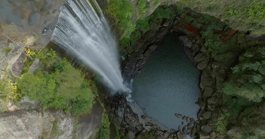 Drone view of a large waterfall dropping into a small pool of water surrounded by rocks at the bottom