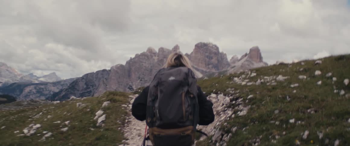 A hiker wearing a backpack is walking up a rock covered mountainside under a cloudy sky