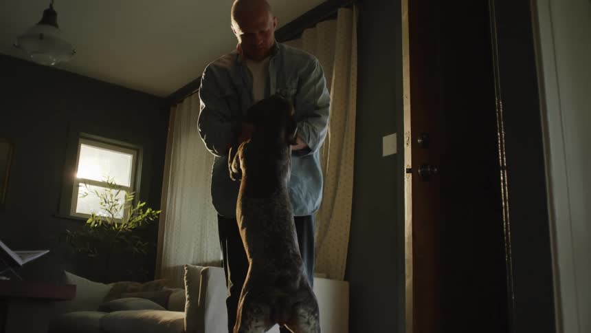 A man walks into his living room and is greeted by his dog as he stops to pet it and the dog walks out the door
