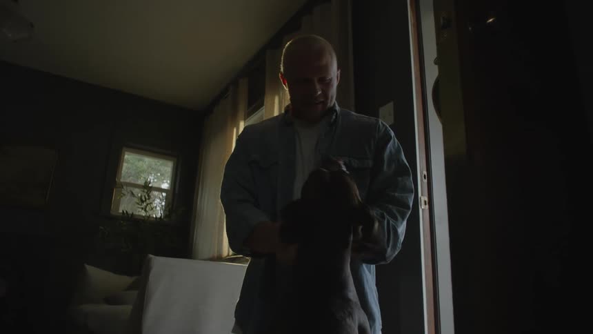 A man standing indoors during the day while petting his dog and greeting him with a smile