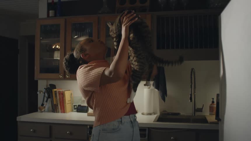 A teenage girl is standing in the kitchen holding her cat as she lifts it up and kisses it