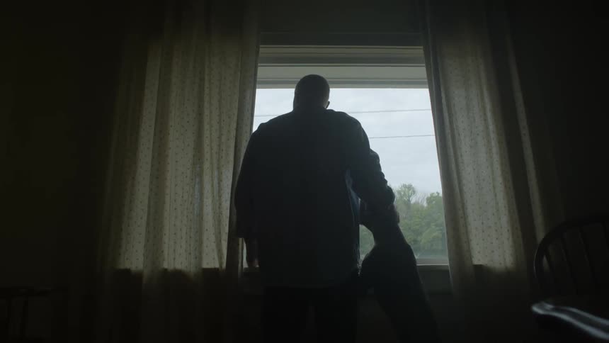 The backside of a man standing in front of a bay window during the day as his dog jumps up and he pets him
