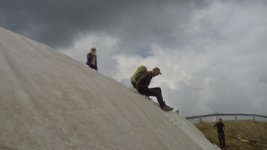 Man with a backpack sliding down the snow on a slope next to two other man watching it outdoors during day time