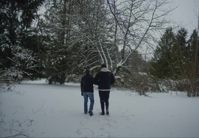 One adult holds the arm of another adult as they walk in the snow near a grove of trees