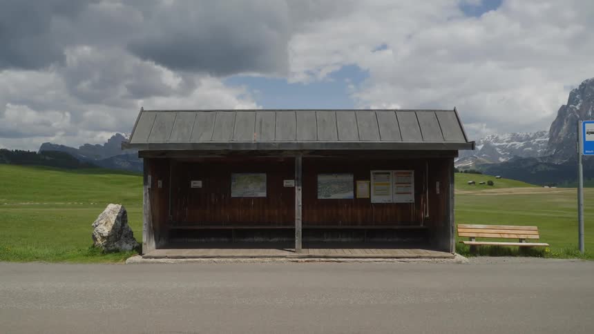 A wooden bench next to a shelter at a rural bus stop with green fields and snow dusted mountains behind