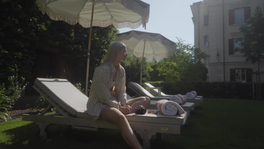 Woman sits outdoors on chaise under umbrella