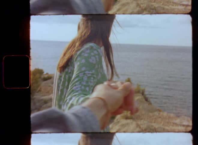 Beautiful brunette woman in green summer dress taking and holding another person’s hand and walking towards a cliff on the seaside or beach leading the way in a romantic love couple moment during a holiday date on vintage 8mm film