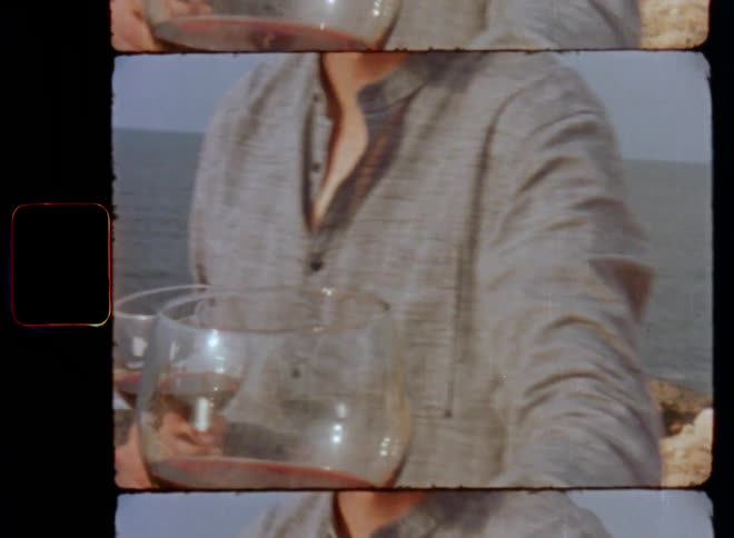 Young man on a cliff by the sea with short beard gray shirt and long hair hands a glass of wine to a person filming in first person perspective raises his hand holding the red wine to say cheers on 8mm film footage with retro vibes