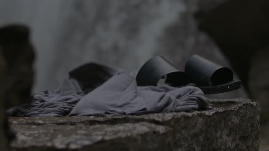 Waterfalls flow down the side of a cliff as a pair of sandals and a shirt fall on a flat rock