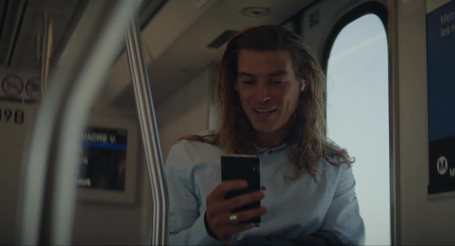 A man with long hair is on a train and is talking on the cell phone with someone and smiling