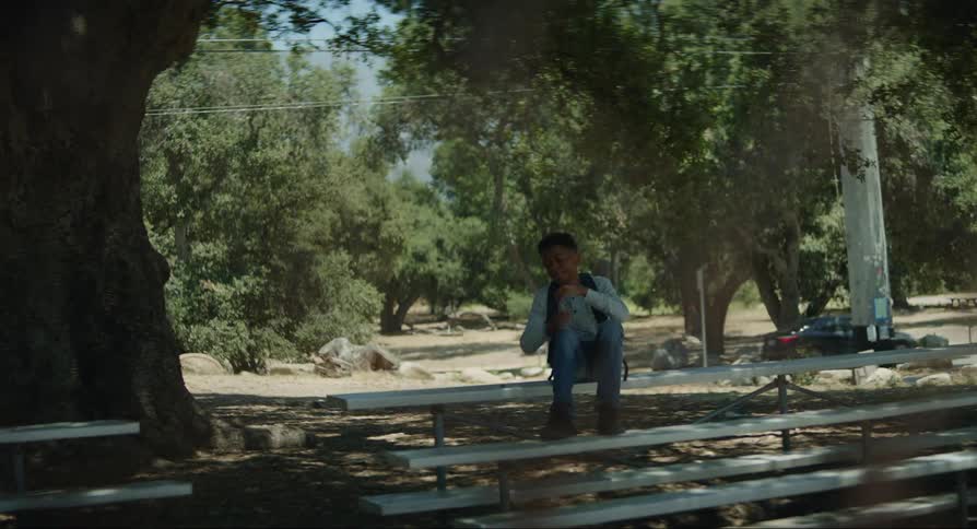 Black teenager boy sitting on a bench outside next to the street and a park seen through a fence