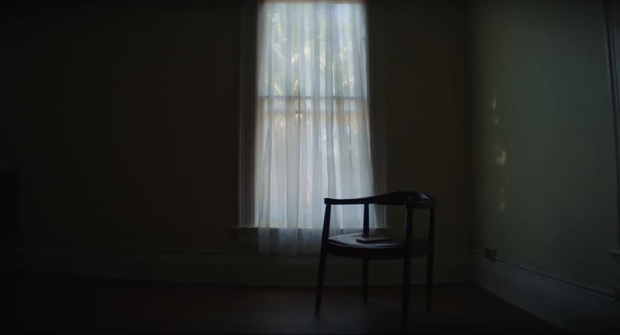 A wood chair in front of a window with a waving curtain in a dark room