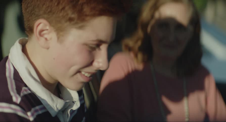 A teenage boy is sitting in the passenger seat of a car in a parking lot talking to his mother
