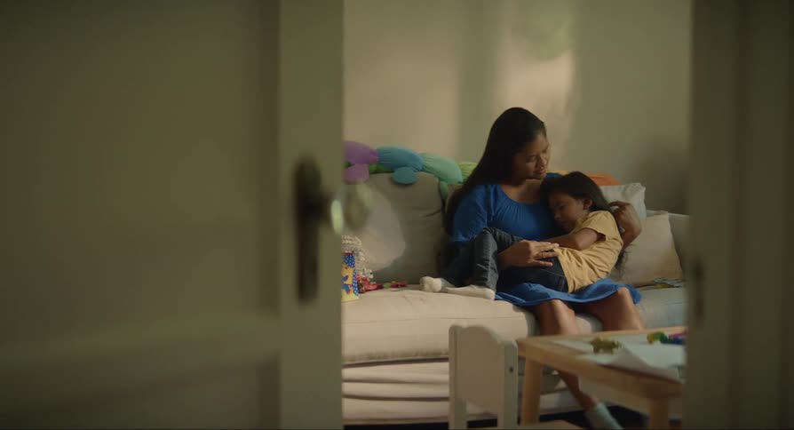 A mother sits on a couch with her daughter in her lap stroking her hair