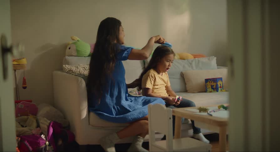 A woman combing the hair of her daughter who is sitting playing with her toy