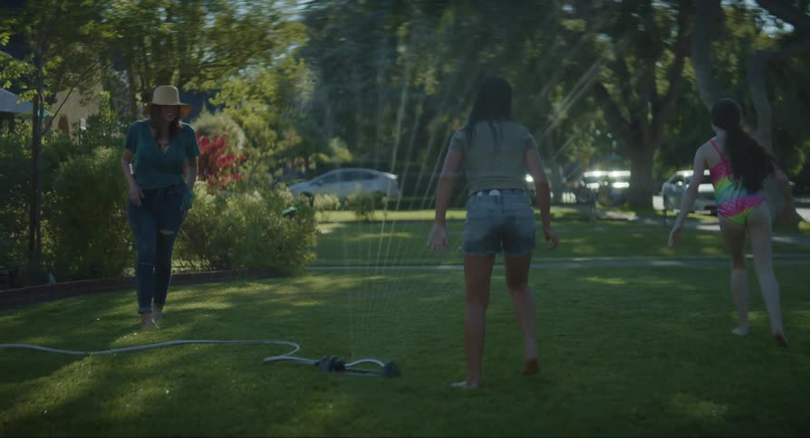 A mother and her daughters are running through a sprinkler in their front yard during the summertime