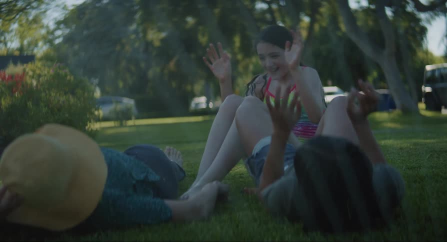Two teenagers and an adult woman lie or sit on a lawn getting sprinkled while playing with each other