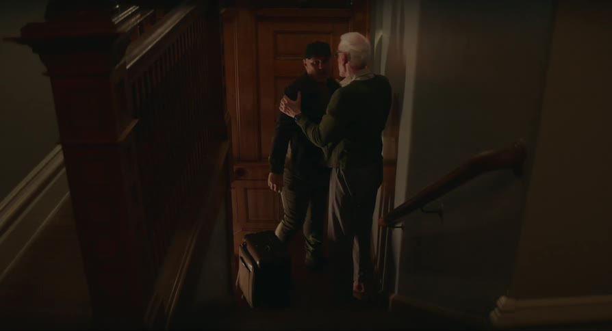 An elderly man comes down the stairs and hugs a man who has arrived at the door with suitcases