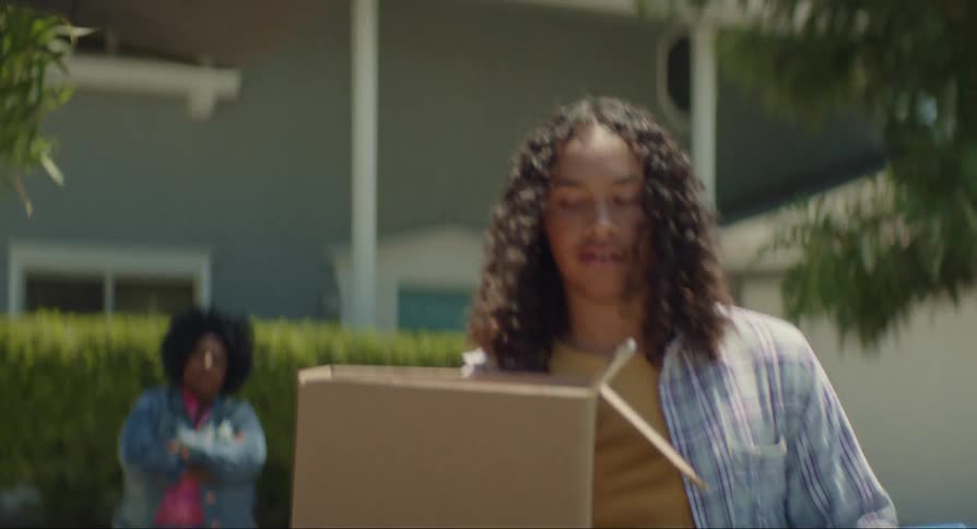 Black woman and a teenager boy kissing and hugging and then the boy carries a cardboard box and a plastic basket into the trunk of a car outdoors during day time