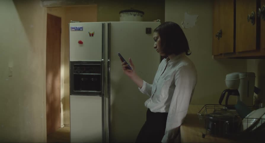 A bored young woman stands in a kitchen scrolling through her smart phone
